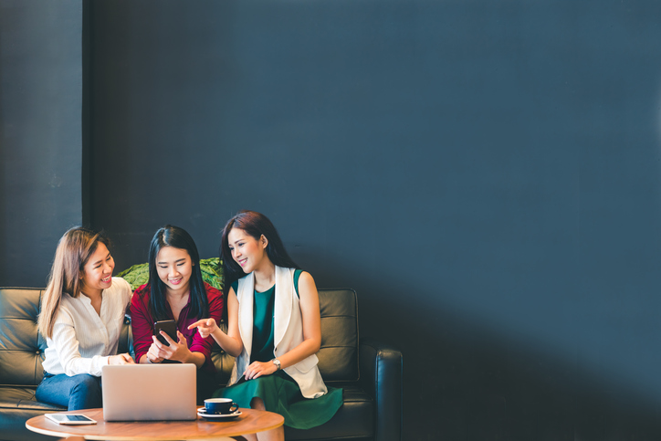 Three beautiful Asian girls using smartphone and laptop, chatting on sofa at cafe with copy space, modern lifestyle with gadget technology or working woman on casual business concept