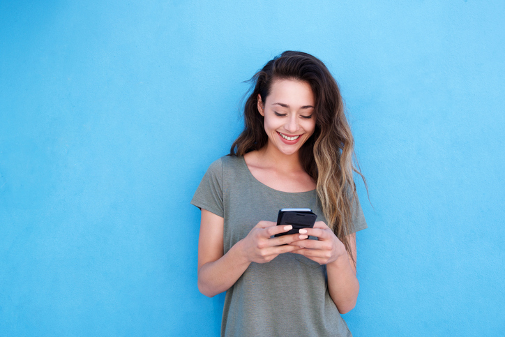 Front portrait of young smiling woman using mobile phone against blue background