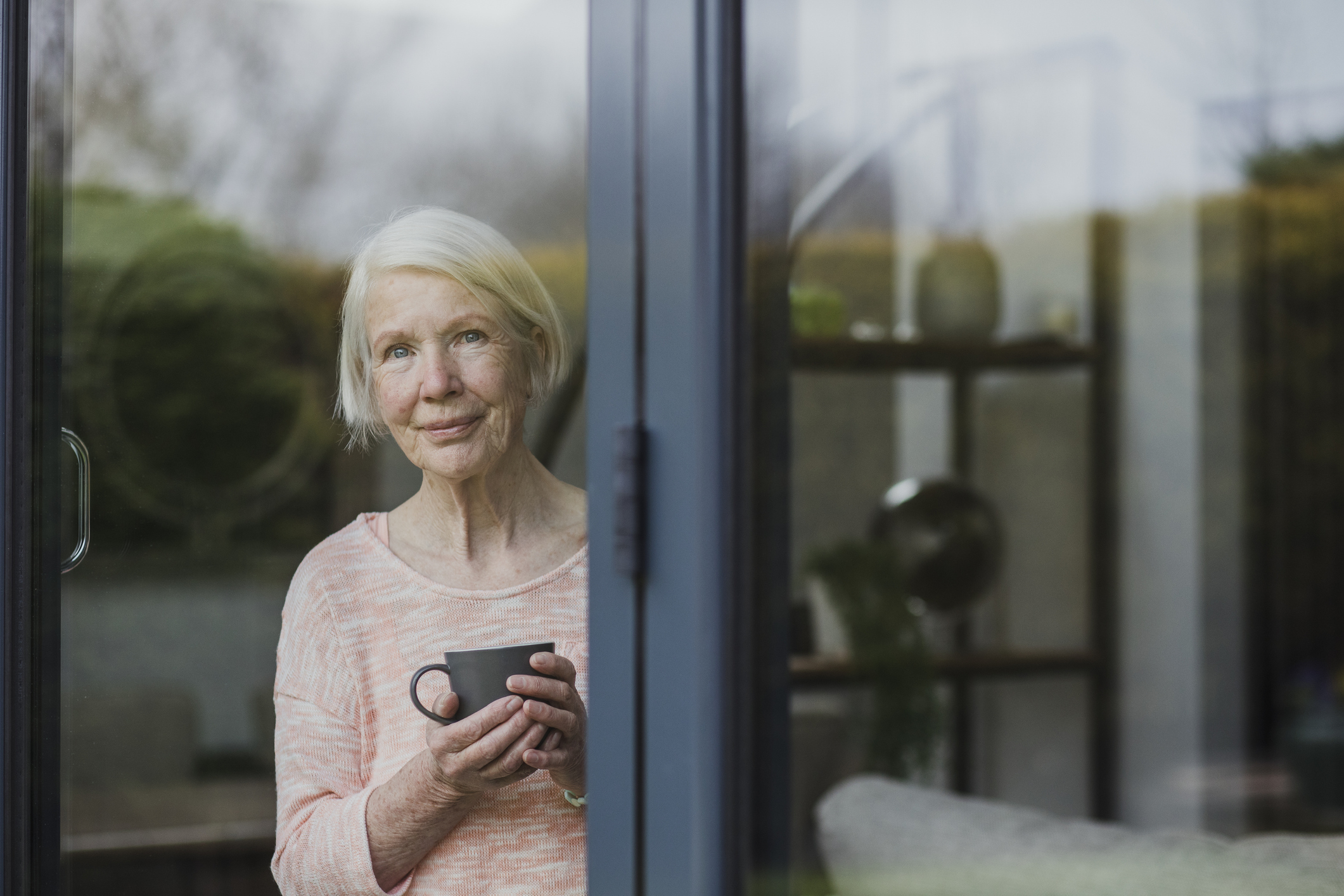 Senior woman is standing at the window in her home, enjoying a cup of tea.