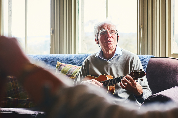 Happy senior man singing song and playing guitar for a woman sitting in front at home