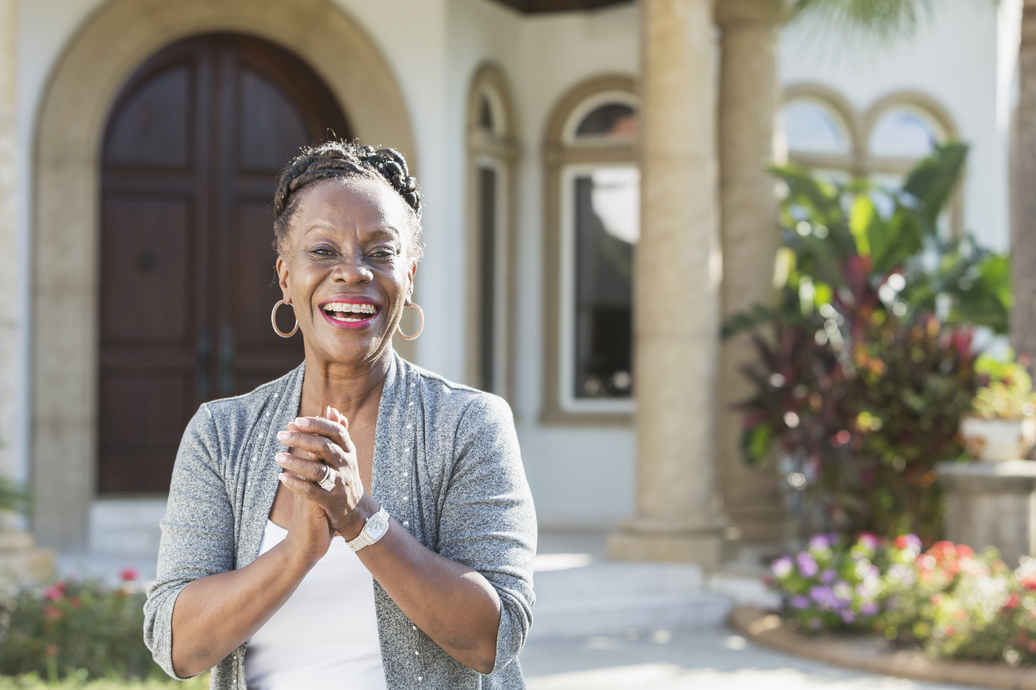 A happy senior African-American woman in her 60s standing outdoors, in front of her home, smiling confidently at the camera, hands clasped.