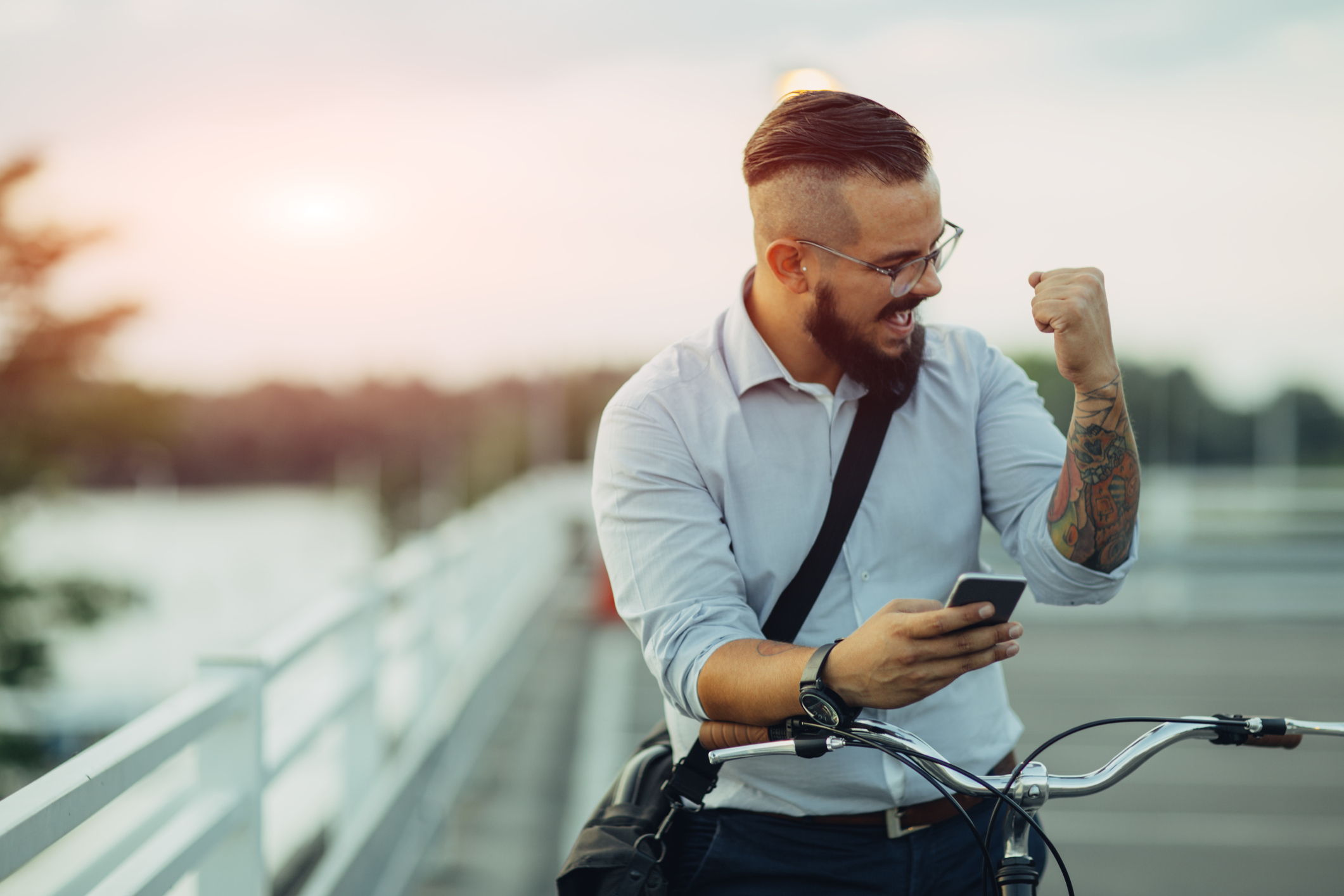 Young Businessman pushing his city bicycle and using his smart phone. He has tattoos and hipster haircut. He just got positive reports from stock market. Using app on his smart phone. He is celebrating his success with fist up.