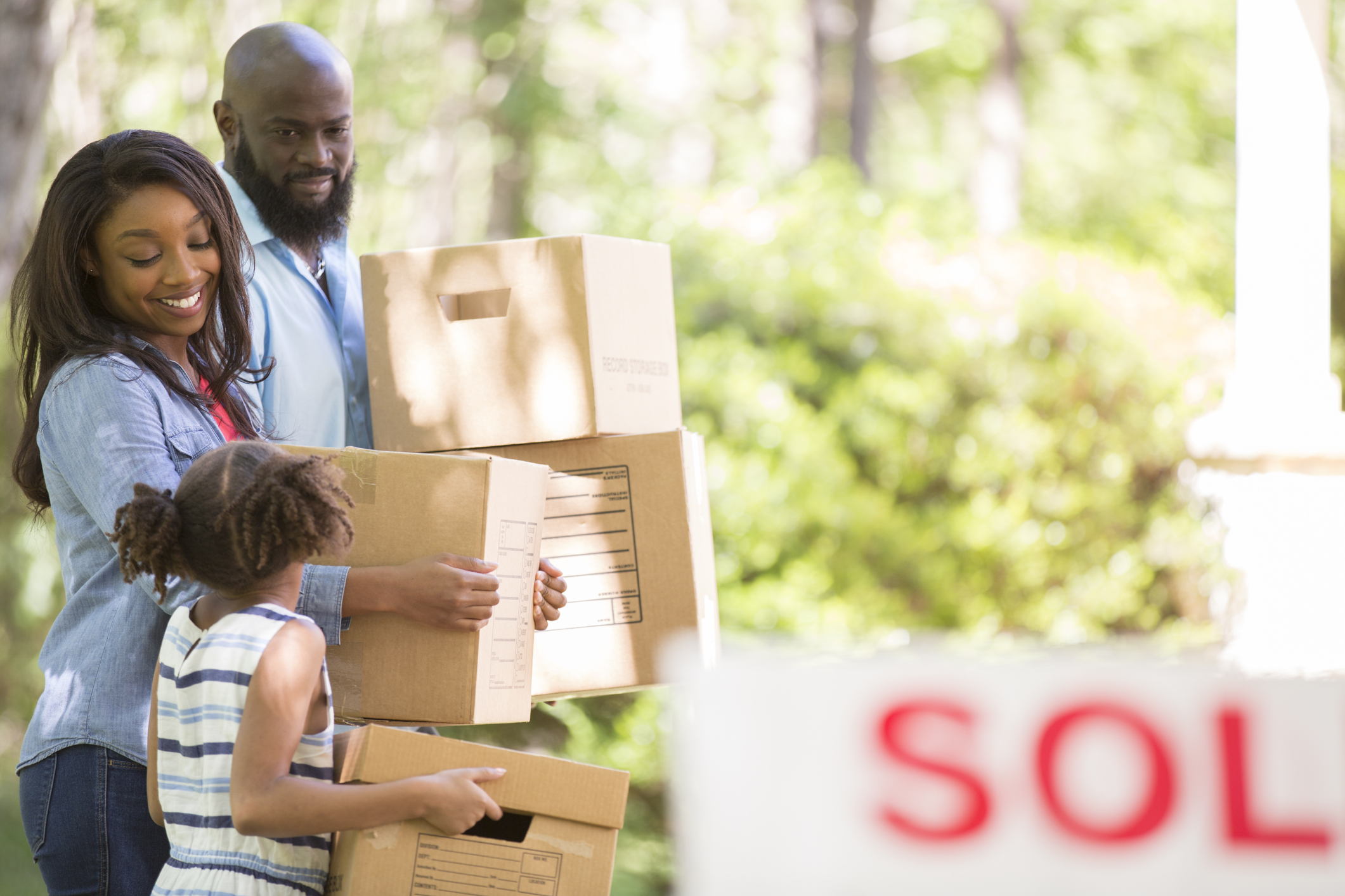 African descent family moving into a new home. Mother, father and daughter. Real estate sign. Home in background. Spring or summer season.