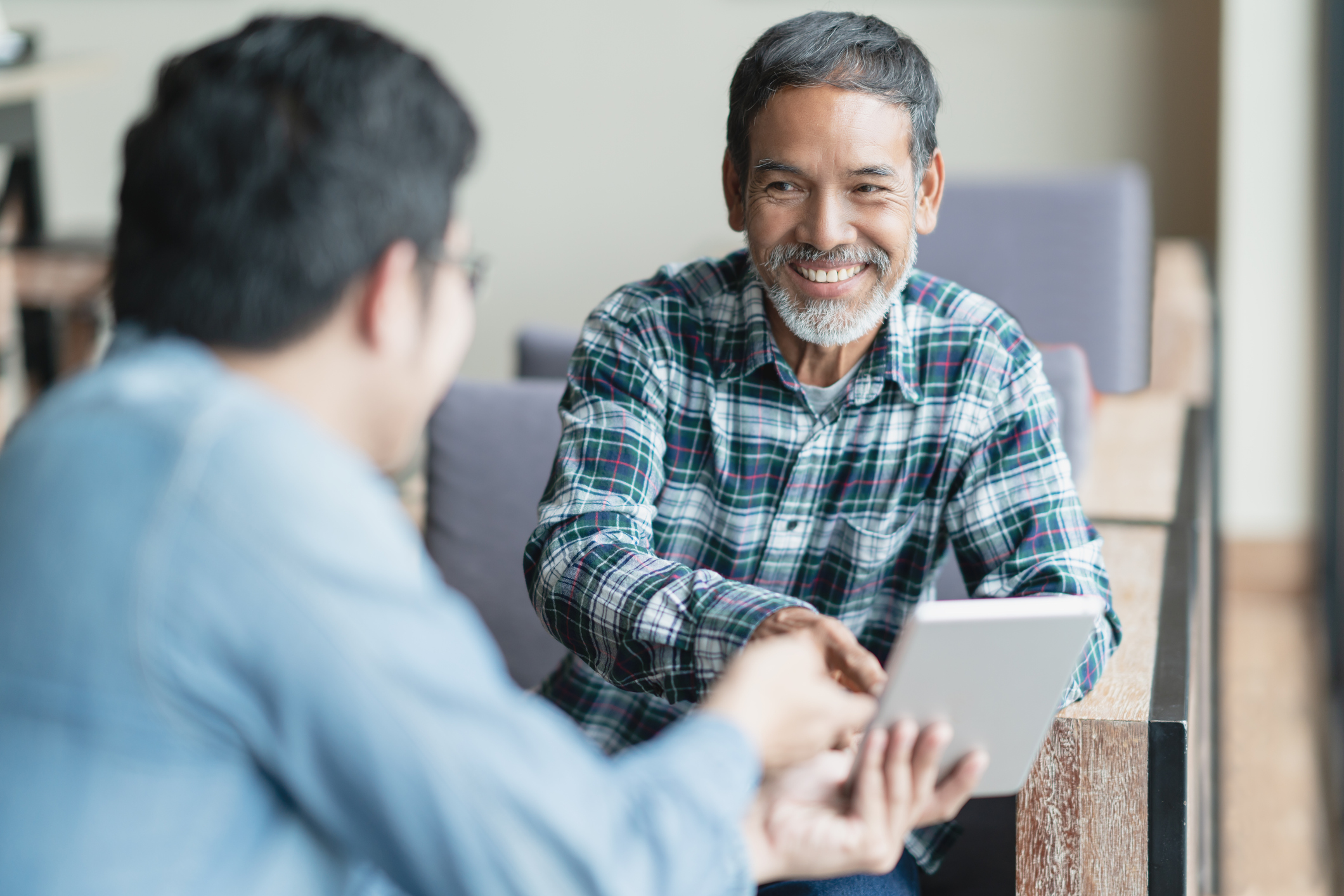 Happy old short beard asian man sitting, smiling and listen to partner that showing presentation on smart digital tablet. Mature man with social media technology teaching or urban lifestyle concept.