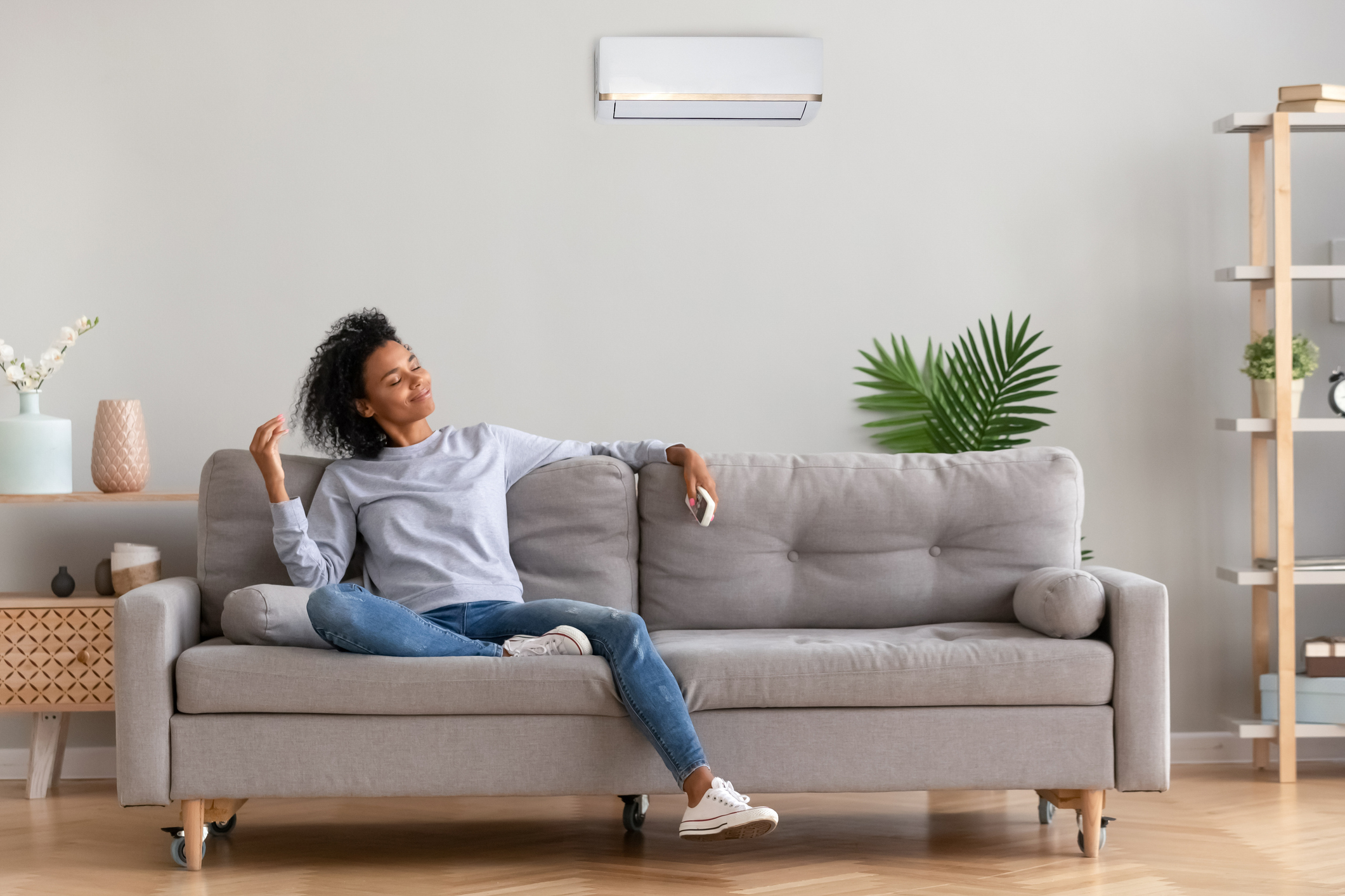 African american relaxed woman sitting on comfortable couch in living room at modern home holds air conditioner remote control enjoying breathing fresh cool air at summer or warm air at winter season