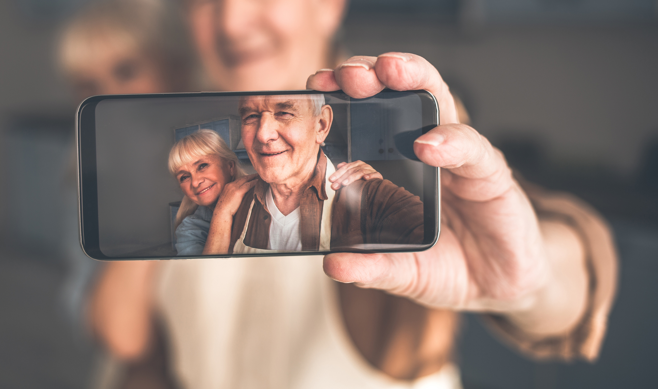 Always happy together. Portrait of glad old loving couple embracing while taking photos. They are standing in kitchen and smiling. Focus on smartphone