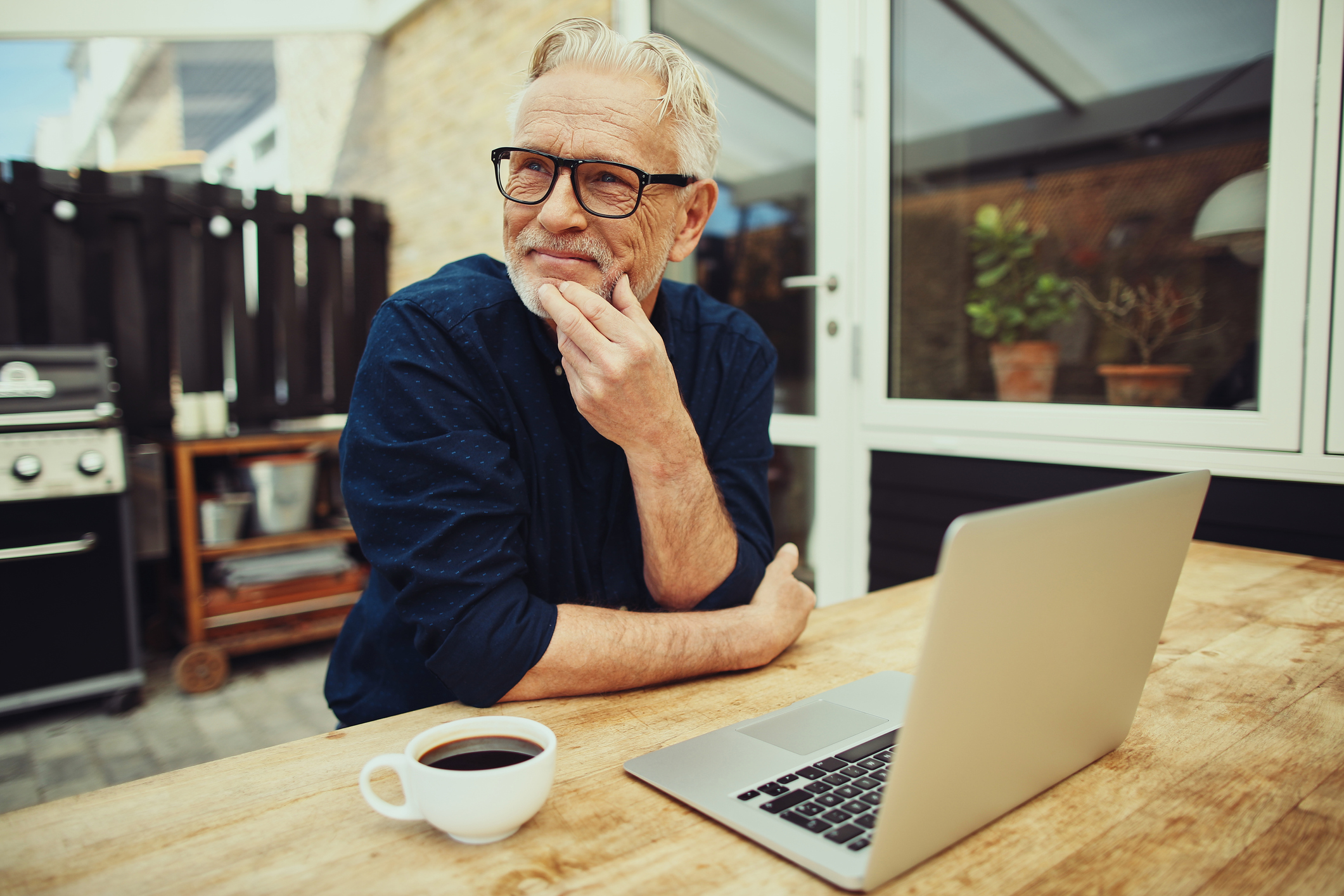 Smiling senior man sitting at a table outside on his patio working online with a laptop and drinking a cup of coffee