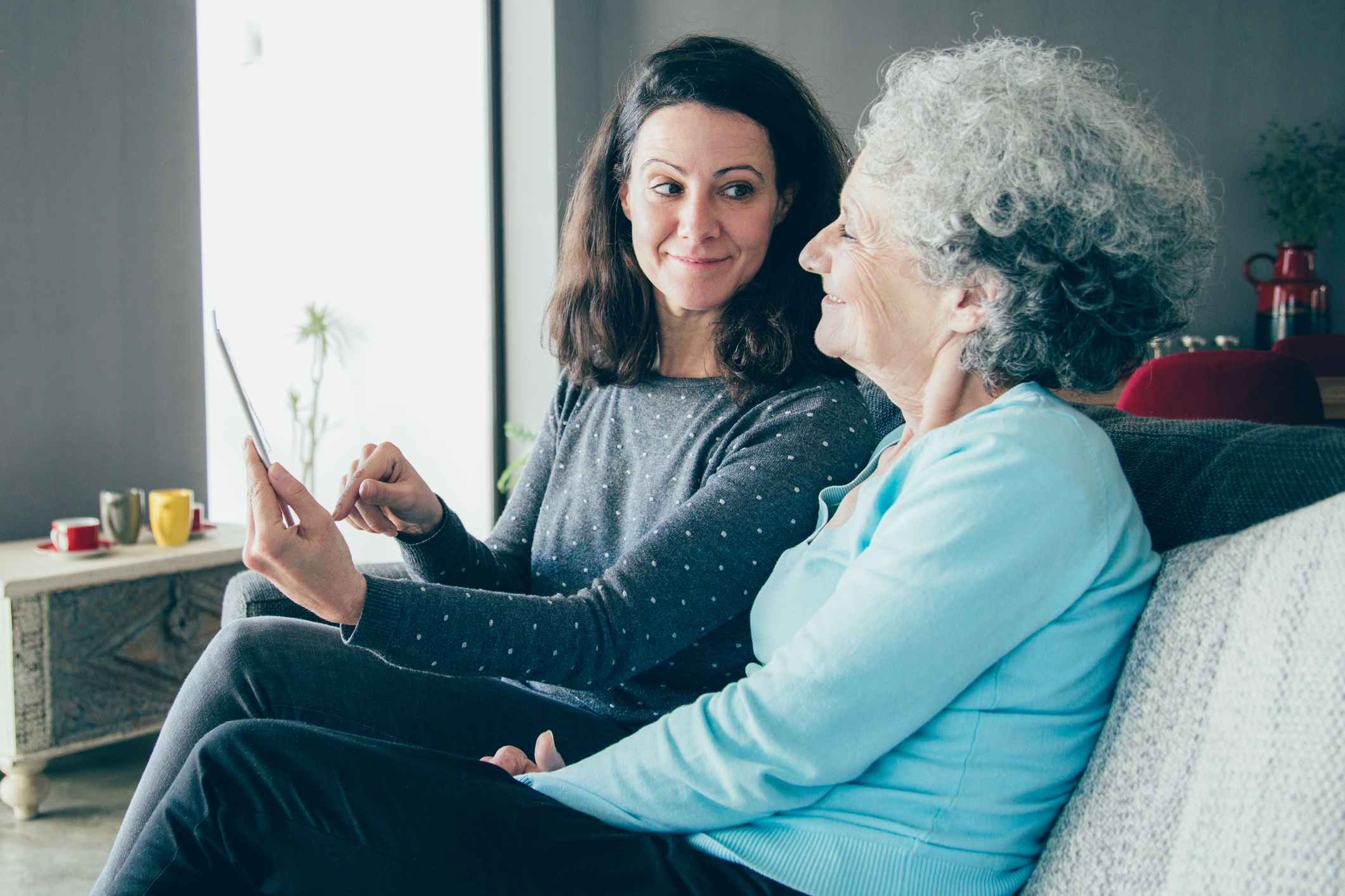 Smiling senior woman and her daughter using tablet computer. Mother and daughter sitting on couch with home interior in background. Technology concept.